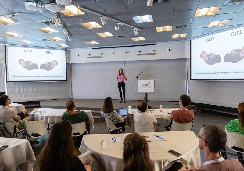 Boglárka egy képernyő előtt prezentál, a közönság hátulról látható ahogy a prezentációt nézi.