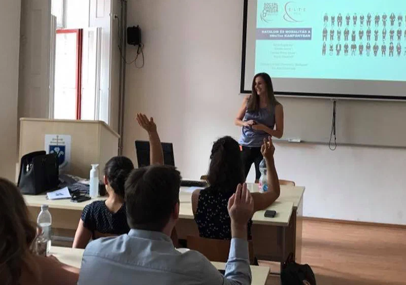 Boglárka egy fehér tábla előtt áll és egy osztálynak prezentál. Néhány diák feltett kézzel ül.