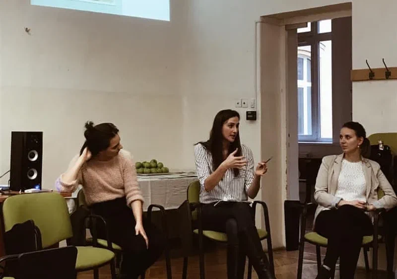 Boglárka és két másik személy egy teremben ül és egy témáról beszélget, a háttérben a falra vetítve egy prezentáció látszik.